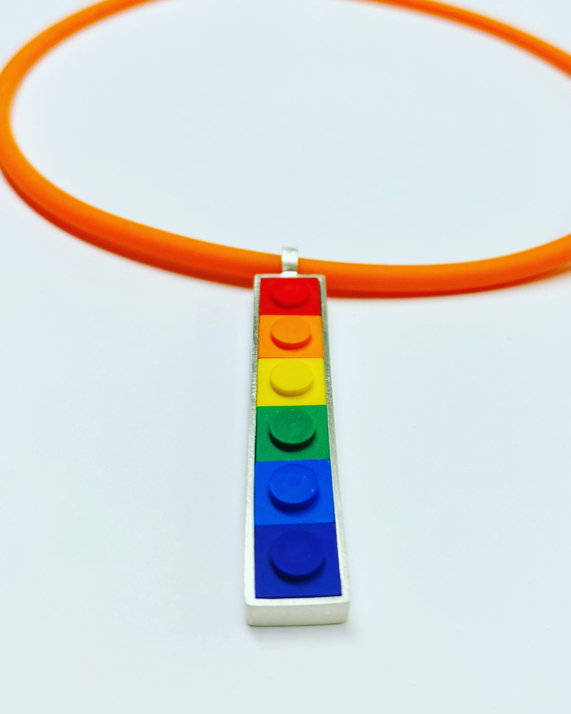 A vertical long pendant that has small square Lego bricks in the color order of a rainbow set in sterling silver, hanging from an orange rubber cord