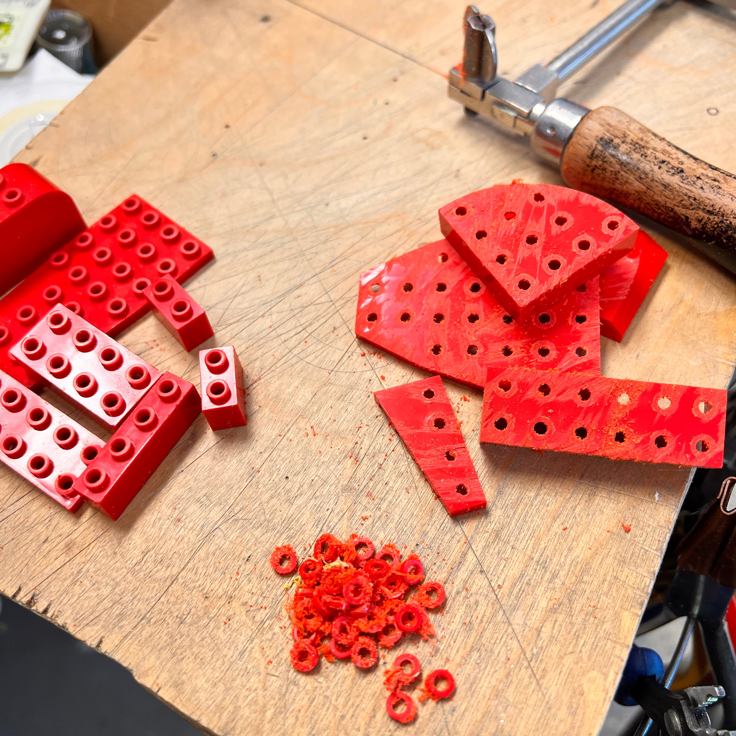 Photo of the process on a work bench showing lego bricks being cut from the top with a jewelers saw to remove the small round tops to create the link for earrings