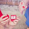 A young boys hands holding an open lego box with a red LEGO dot and diamond ring inside presenting this gift to a geek chic girl