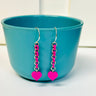 Earrings are displayed hanging off a cup. The earrings have silver jump rings alternating with loops made from cutting off the top studs of pieces of LEGO bricks. LEGO dot heart dangles from the bottom