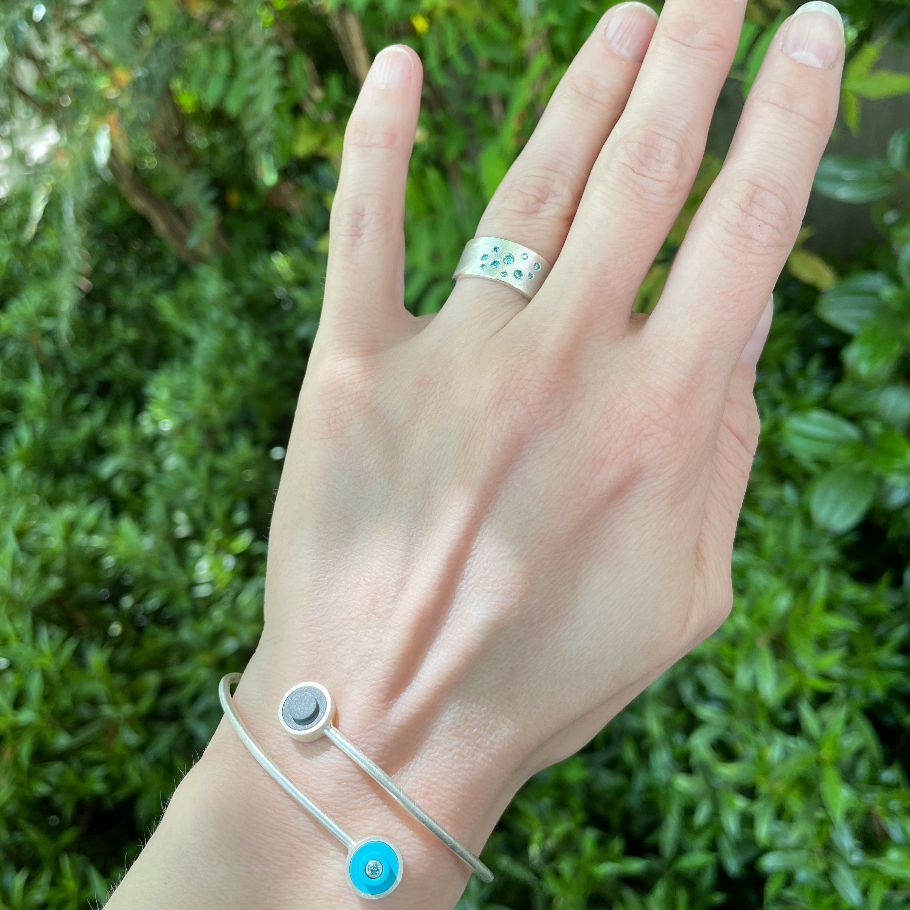 a ladies arm displaying a sterling silver bracelet that wraps around the wrist, has two LEGO dots with diamnds set into them. Finger has a wide band with flush set aqua diamonds