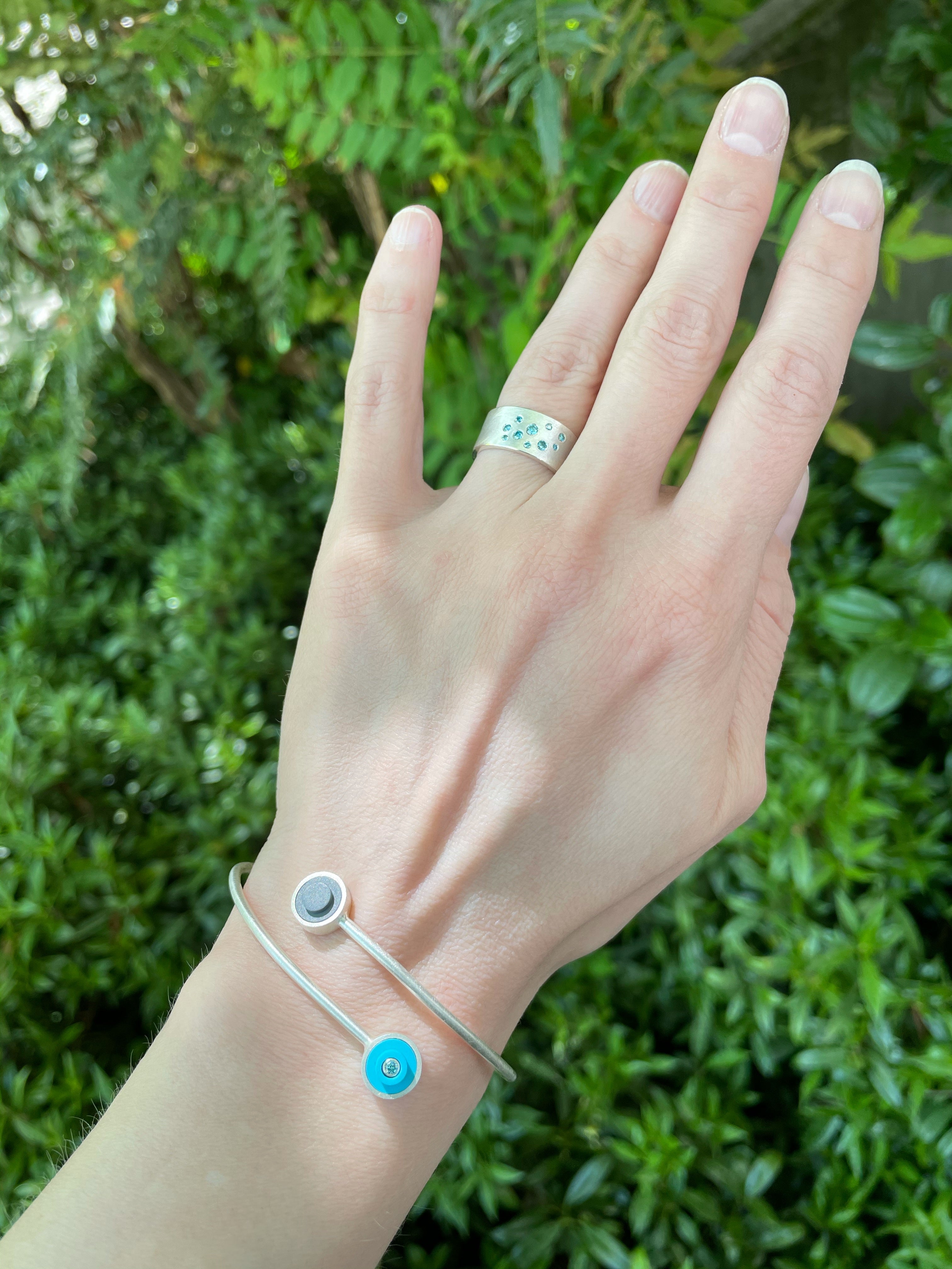 a ladies arm displaying a sterling silver bracelet that wraps around the wrist, has two LEGO dots with diamnds set into them. Finger has a wide band with flush set aqua diamonds