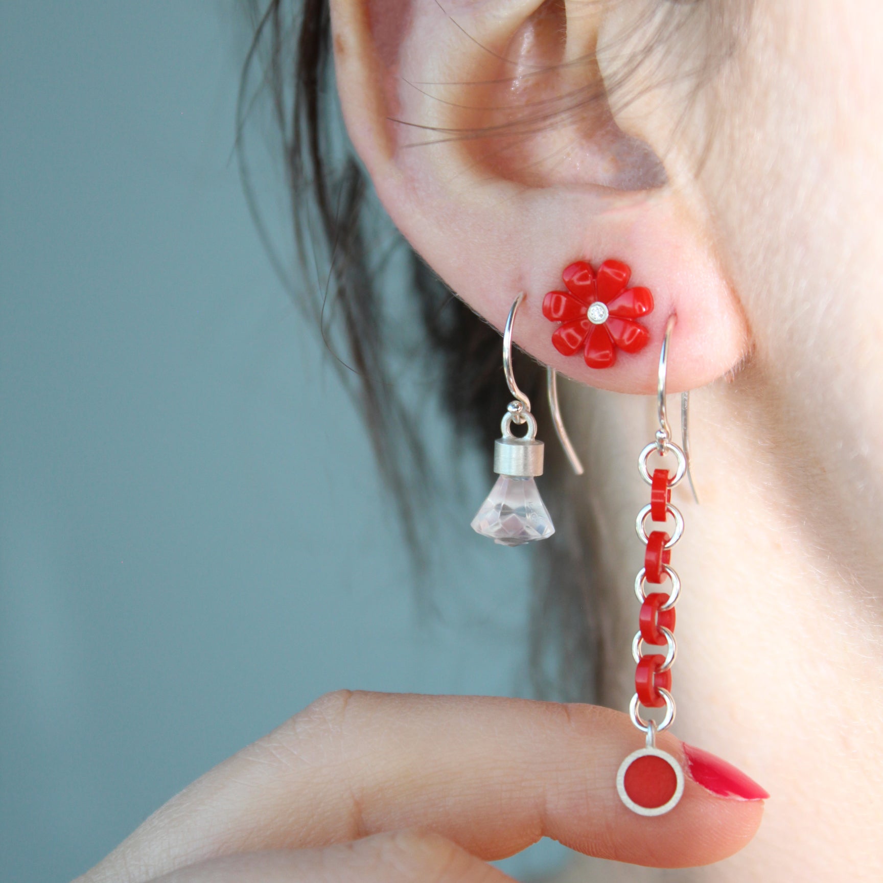 3 earrings shown on a women's ear. First one is dangle with red links, second one is a red daisy stud  with a diamond in the center, third one is the briolette shaped dangle