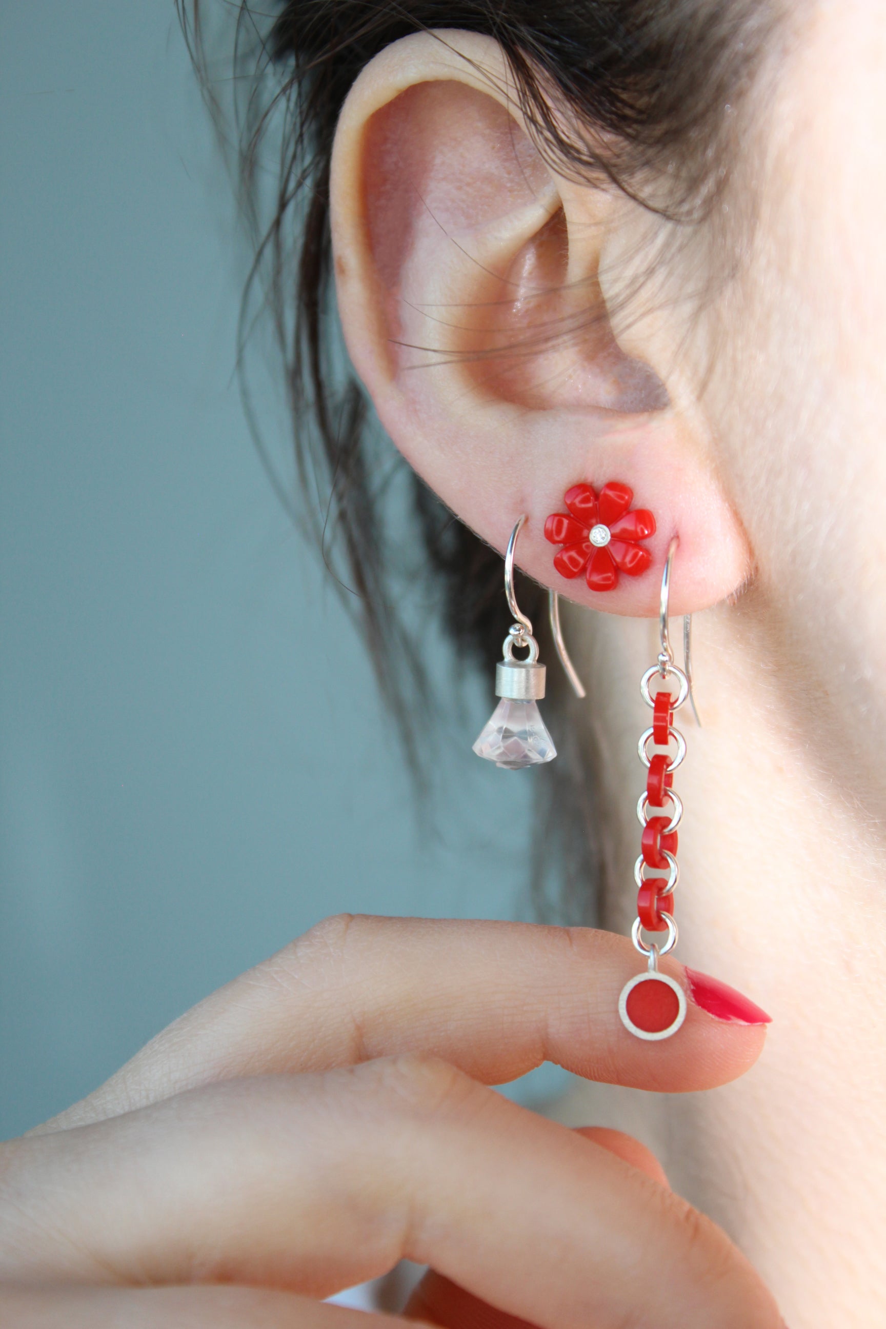 3 earrings shown on a women's ear. First one is dangle with red links, second one is a red daisy stud  with a diamond in the center, third one is the briolette shaped dangle