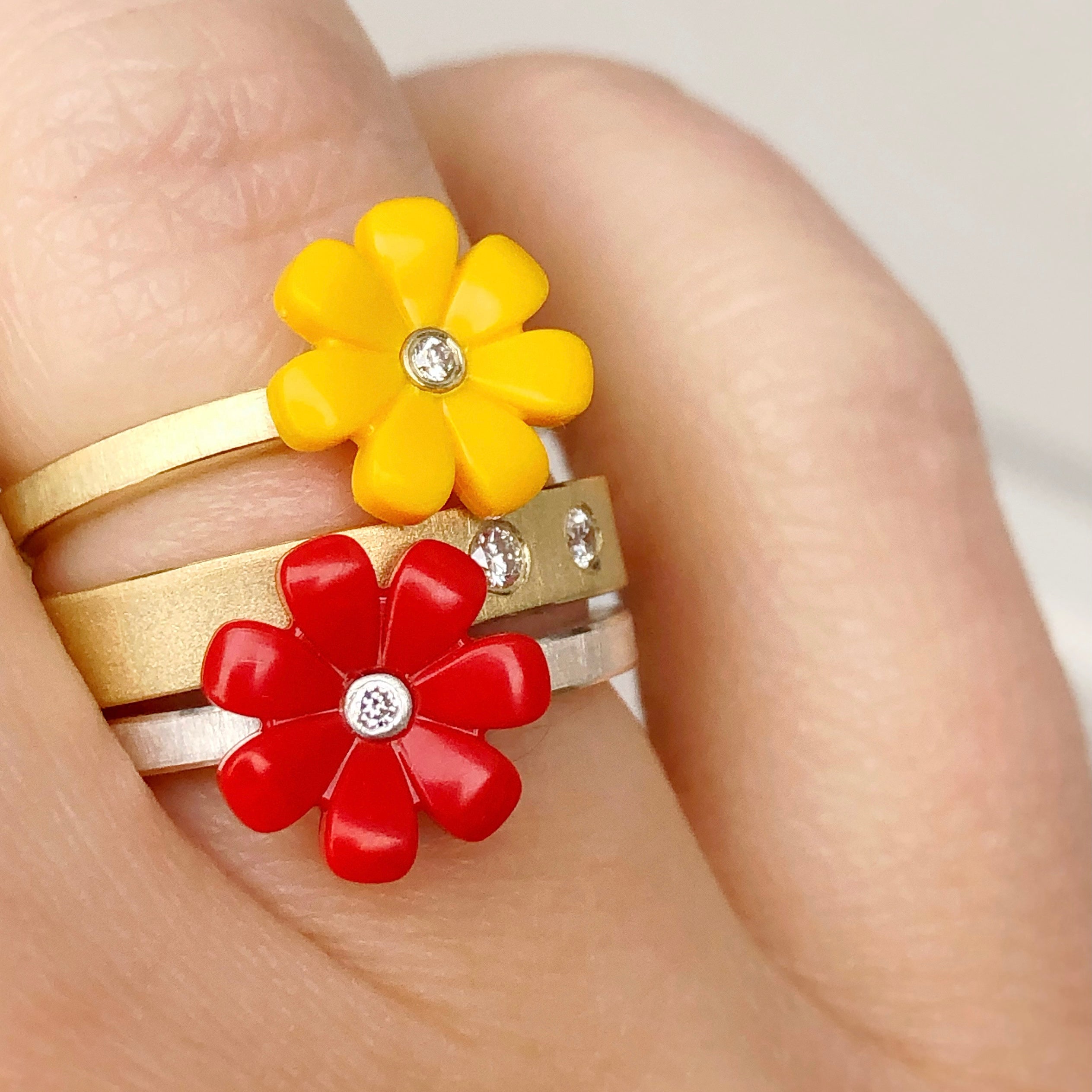 3 18k yellow gold rings shown stacked on a womens finger.  two rings are little daisys, one red and one yellow with a diamond in the center of them. The other ring is a yellow gold diamond band with two diamonds flush set.