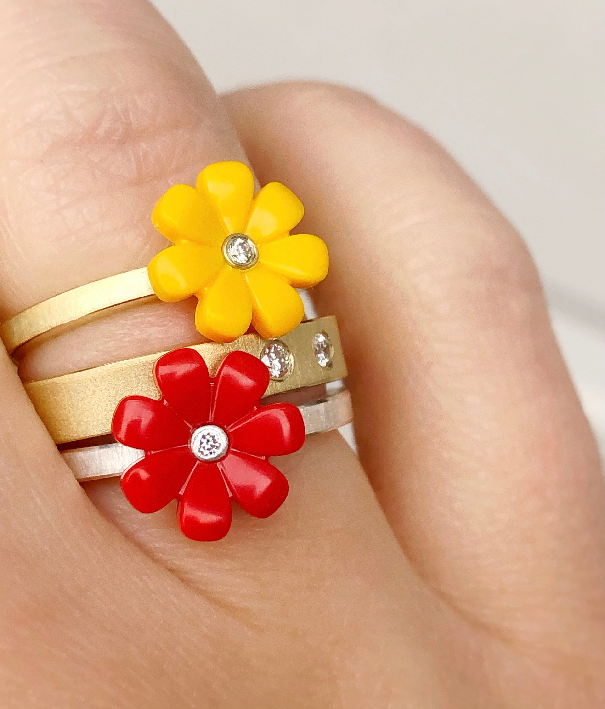 3 18k yellow gold rings shown stacked on a womens finger.  two rings are little daisys, one red and one yellow with a diamond in the center of them. The other ring is a yellow gold diamond band with two diamonds flush set.