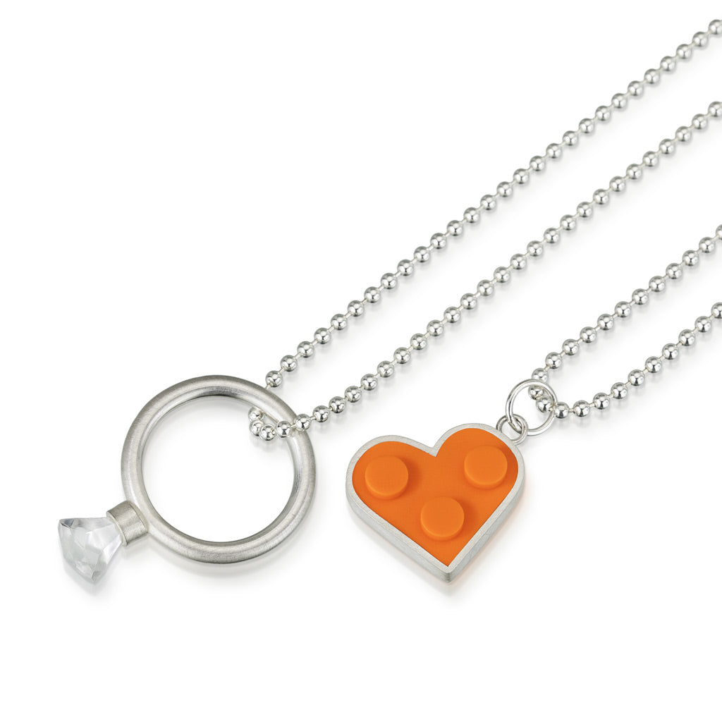 a photo of two sterling silver necklaces. One chain has a sterling silver ring with rounded band with LEGO gem on top. Other chain has a n orange LEGO shaped heart set into a sterling silver.