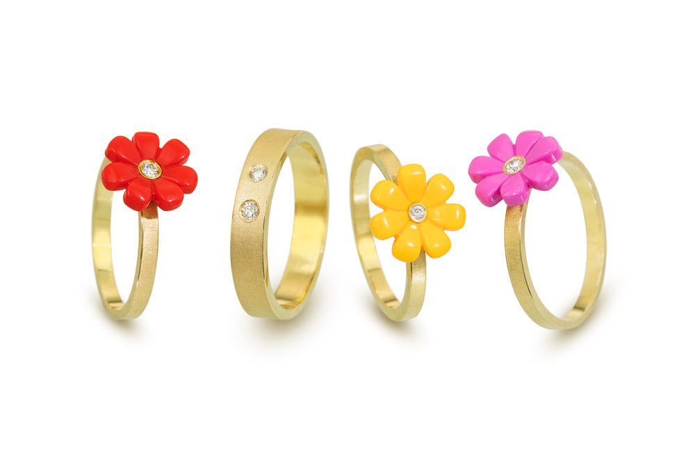 4 18K yellow gold rings in a row standing up. 3 rings have colored daisies. Pink, red , and yellow. Each daisy has a diamond in the center. The other ring is a band with 2 diamonds flush set into the metal
