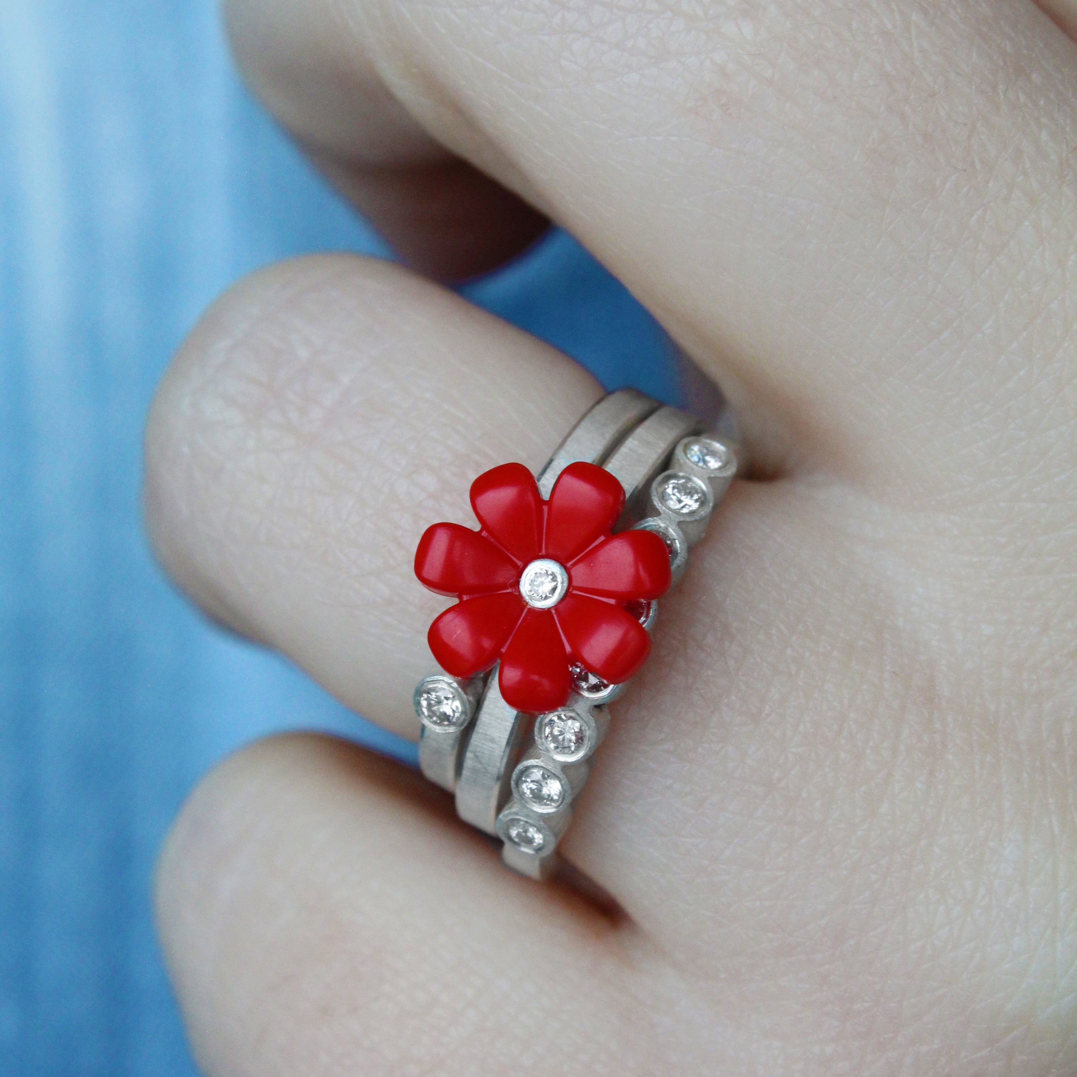 women displaying 3 sterling silver rings stacked on her finger. One has a red daisy with diamond in center, one is a band   sterling silver stackith one diamond set in a bezel and the other one has 8 diamonds going across on the top