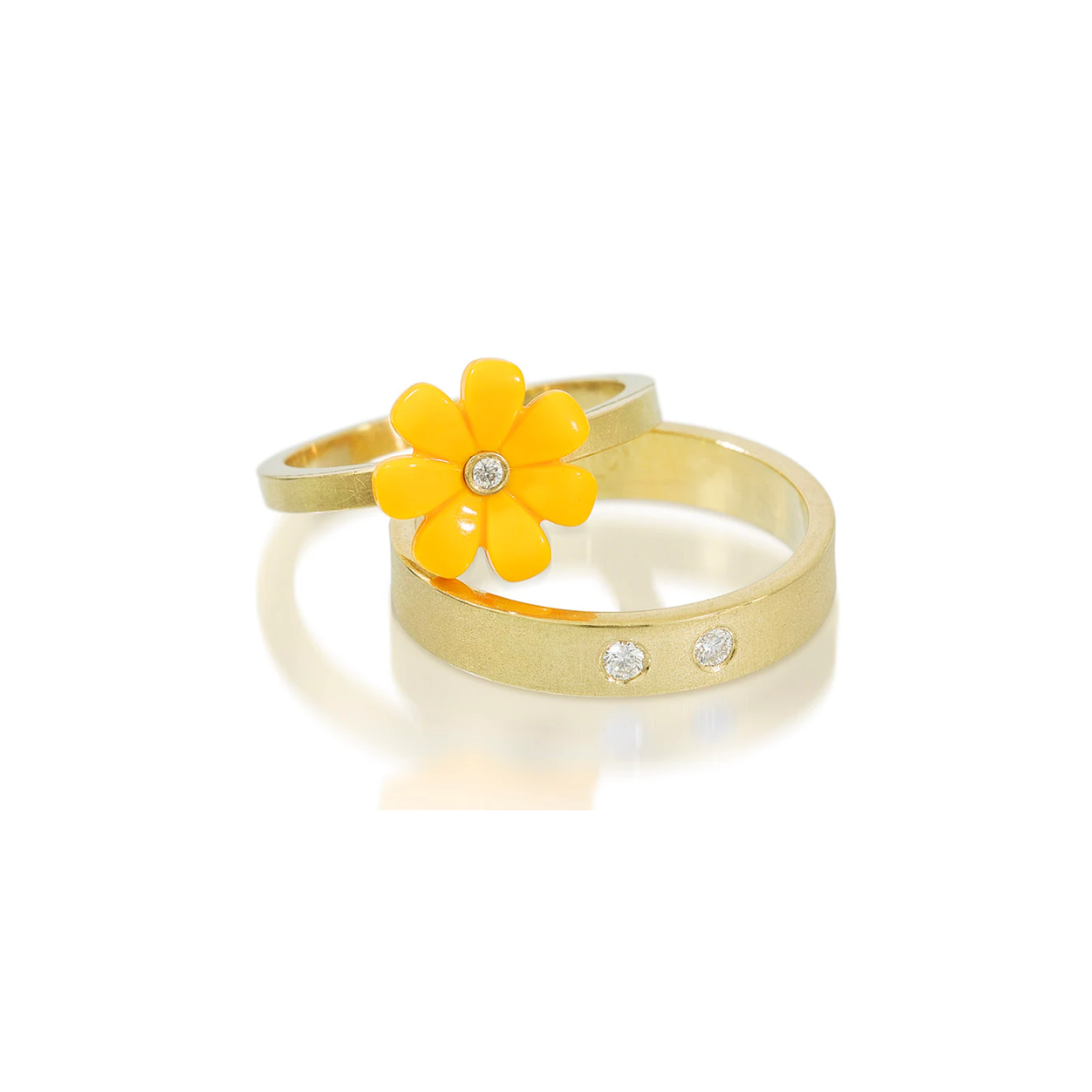 Gold ring with a yellow flower and small diamond leaning up against a gold band with 2 round diamonds on a white background 