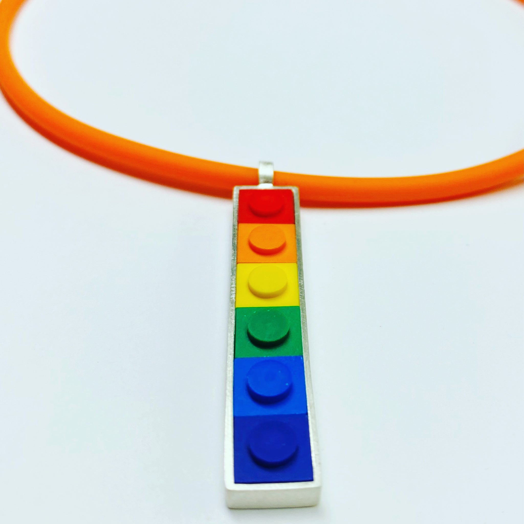 A vertical long pendant that has small square Lego bricks in the color order of a rainbow set in sterling silver, hanging from an orange rubber cord 