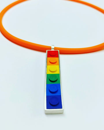 A vertical long pendant that has small square Lego bricks in the color order of a rainbow set in sterling silver, hanging from an orange rubber cord 