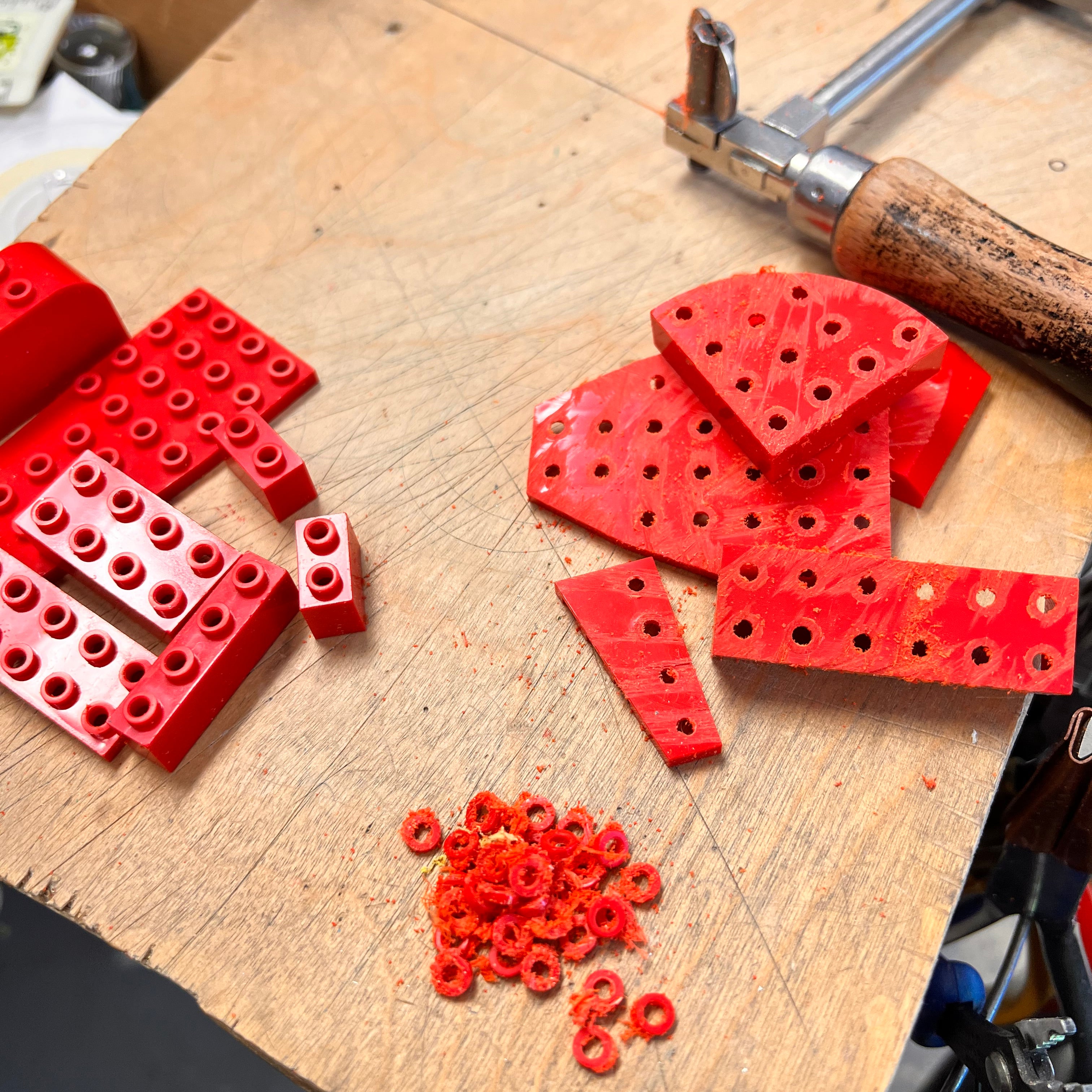 Photo of the process on a work bench showing lego bricks being cut from the top with a jewelers saw to remove the small round tops to create the link for earrings 