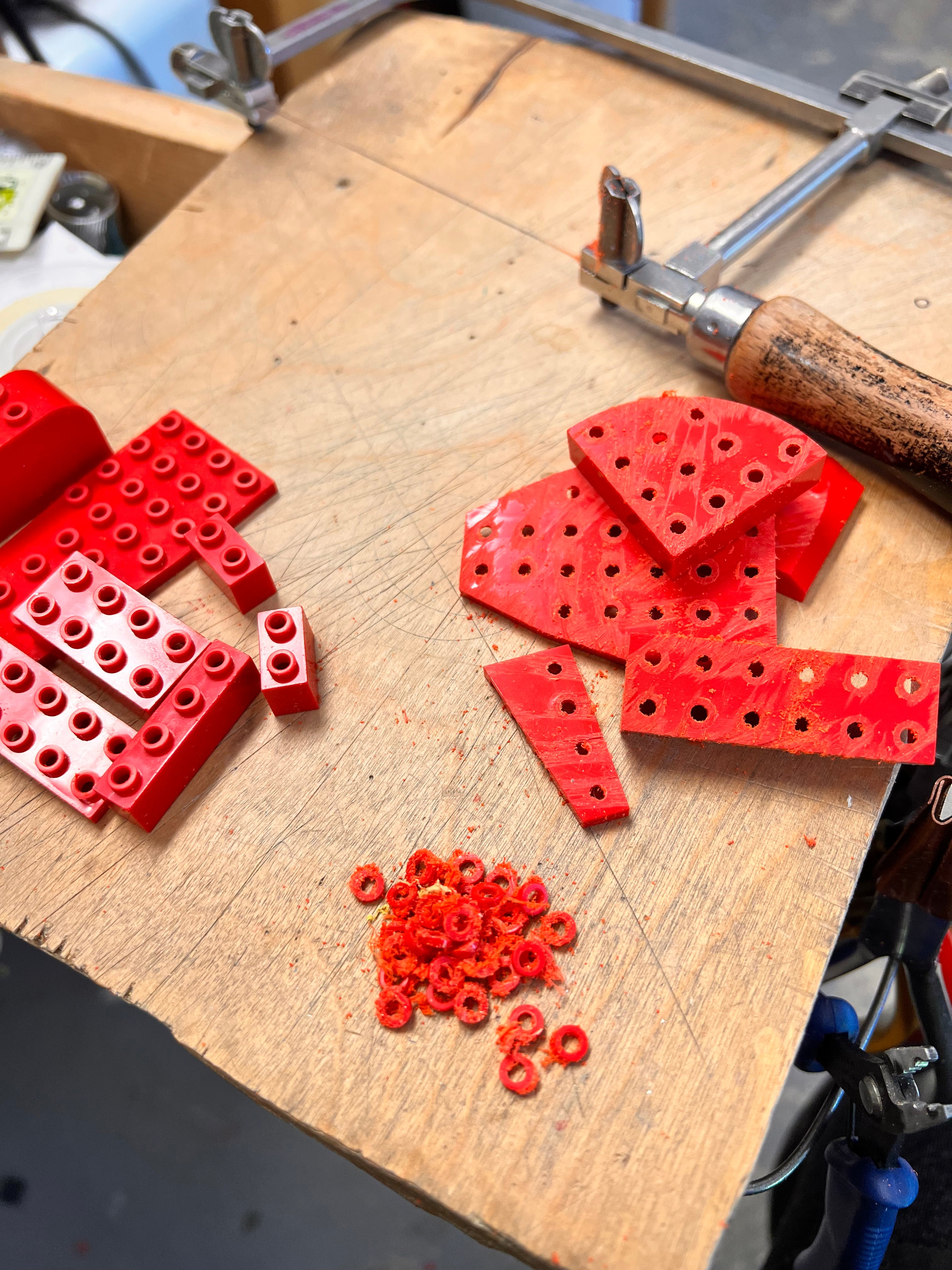 Photo of the process on a work bench showing lego bricks being cut from the top with a jewelers saw to remove the small round tops to create the link for earrings 