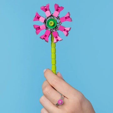 Hand wearing a LEGO and diamond ring holding a colorful LEGO windmill flower against a blue background