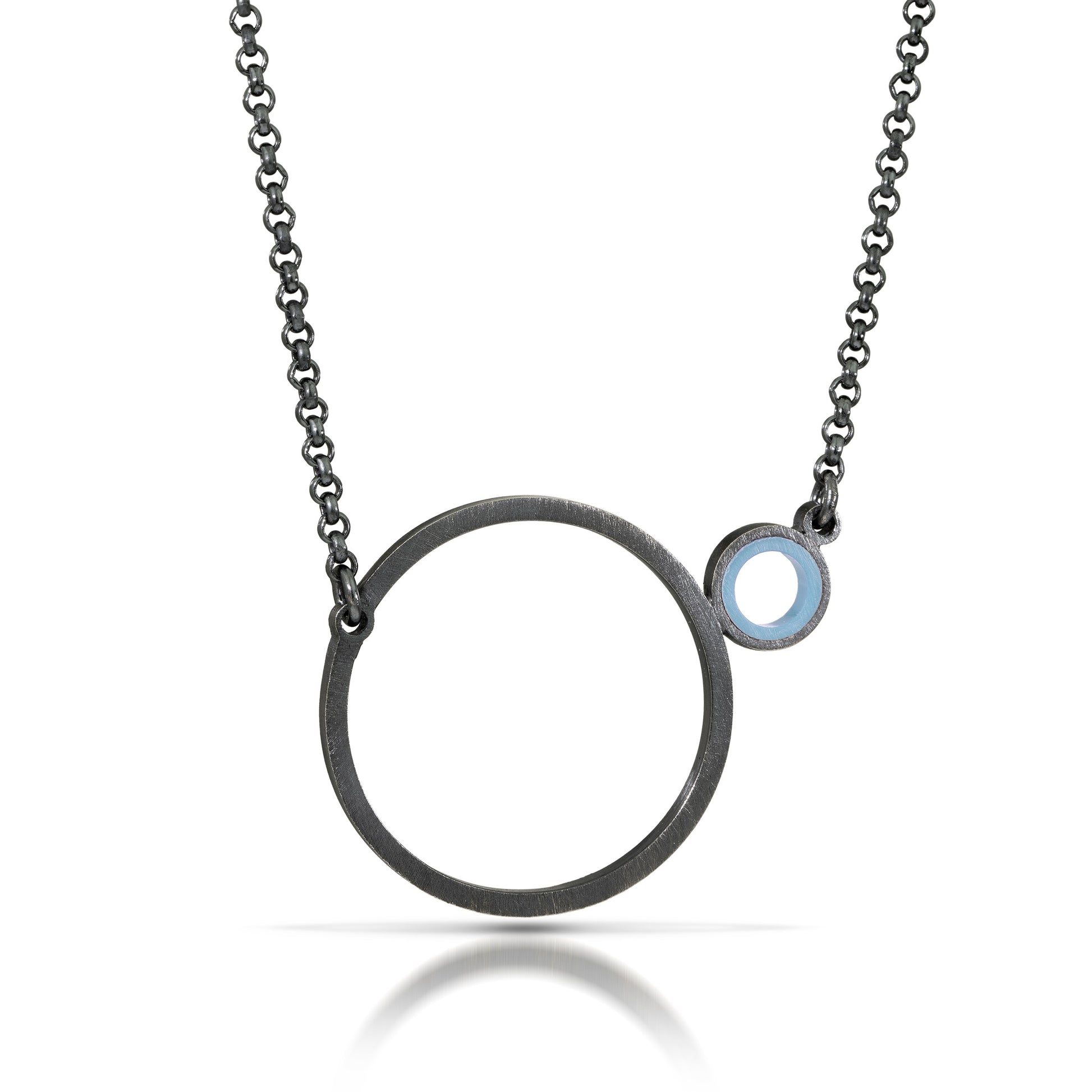 a necklace with a dark finish on the metal with a large circle pedant in center with a small circle attached to the side with a light blue recycled piece of a lego tube inside the small circle