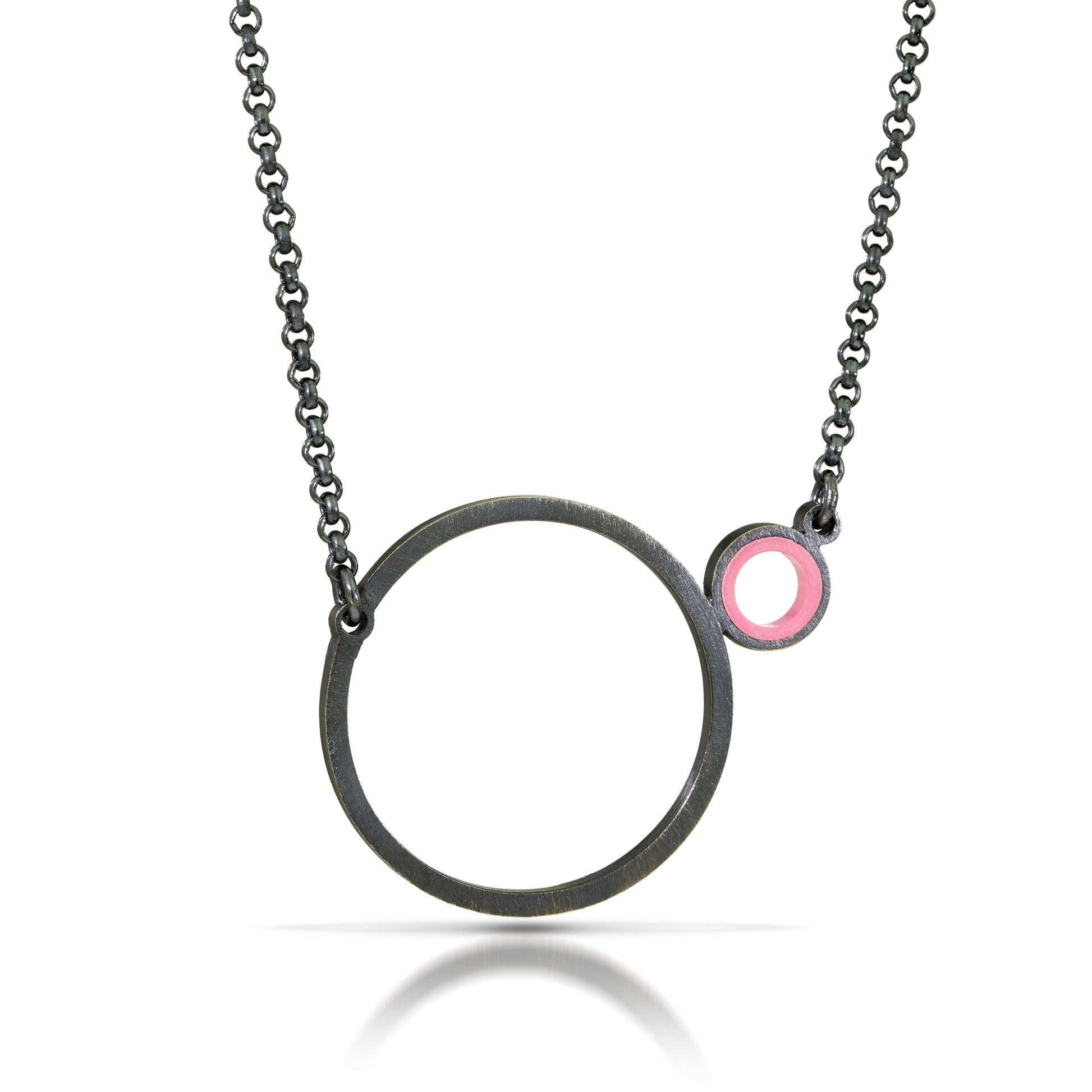 a necklace with a dark finish on the metal with a large circle pedant in center with a small circle attached to the side with a light pink recycled piece of a lego tube inside the small circle