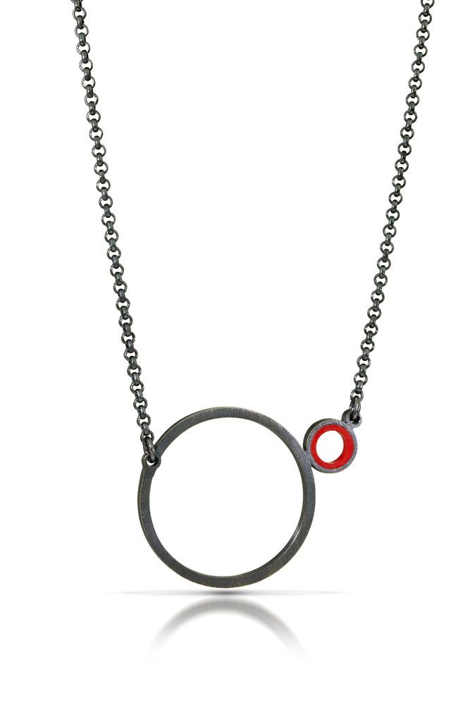 a necklace with a dark finish on the metal with a large circle pedant in center with a small circle attached to the side with a red recycled piece of a lego tube inside the small circle