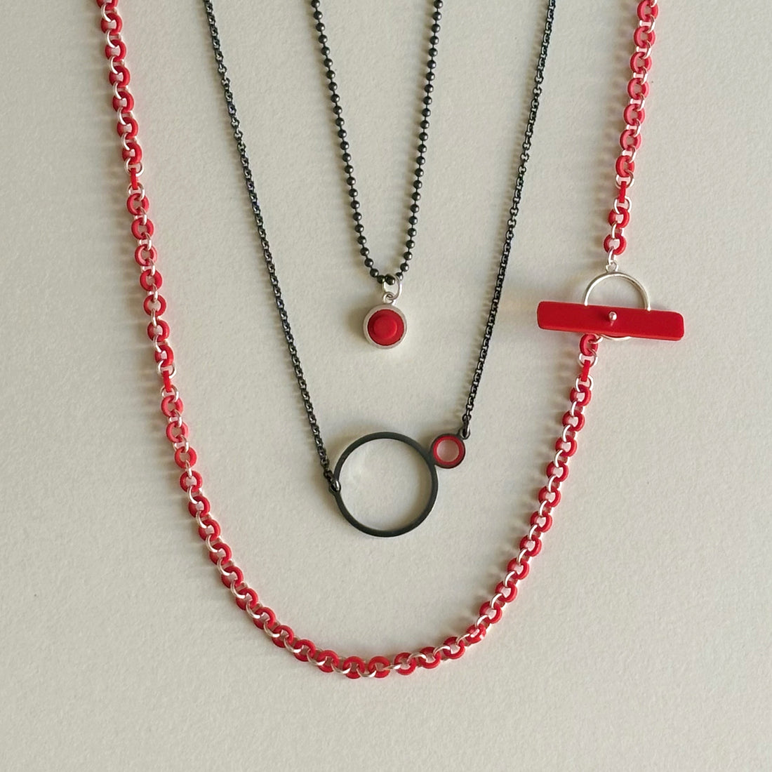 a necklace with a dark finish on the metal with a large circle pedant in center with a small circle attached to the side with a red recycled piece of a lego tube inside the small circle