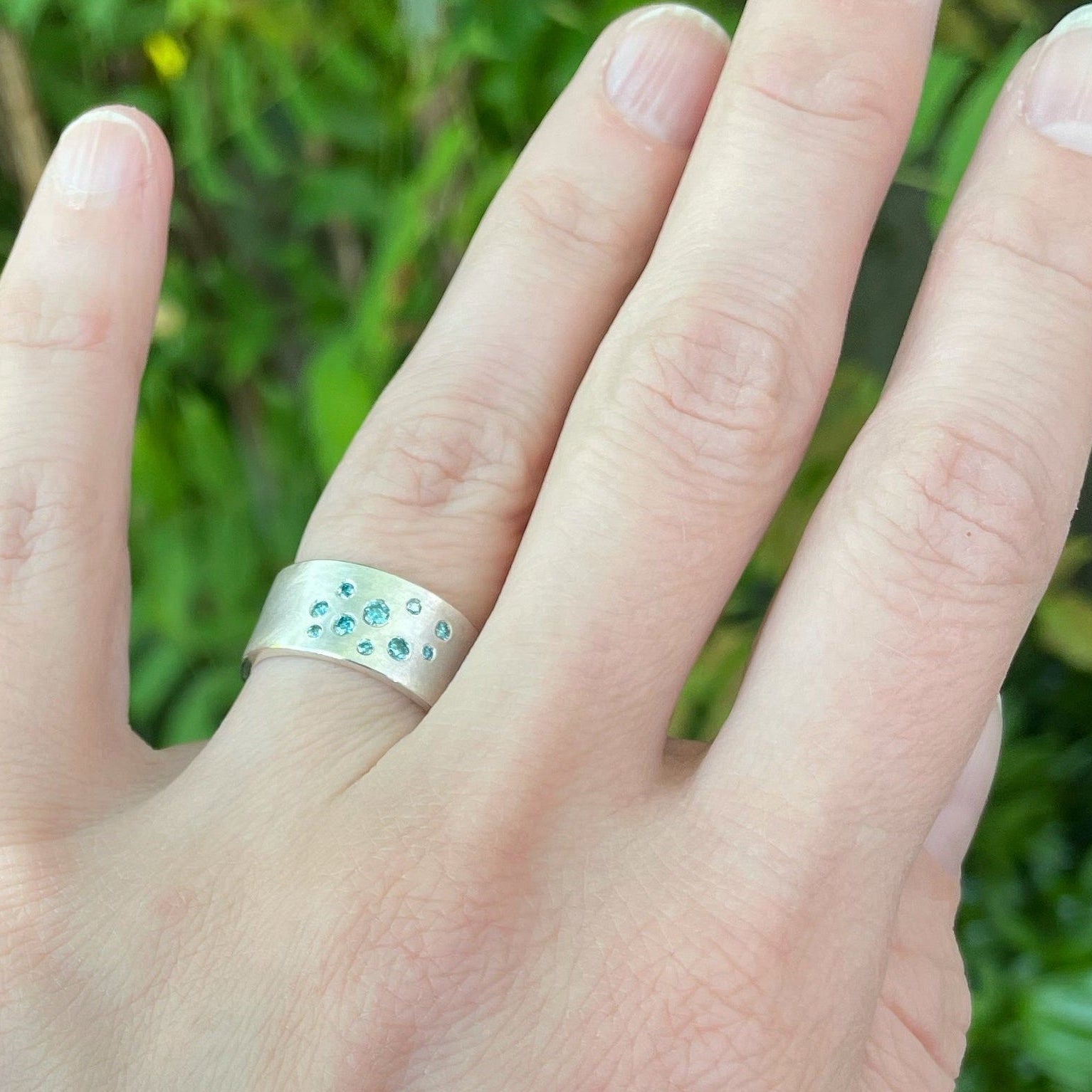 a ladies arm displaying a sterling silver bracelet that wraps around the wrist, has two LEGO dots with diamnds set into them. Finger has a wide band with flush set aqua diamonds 
