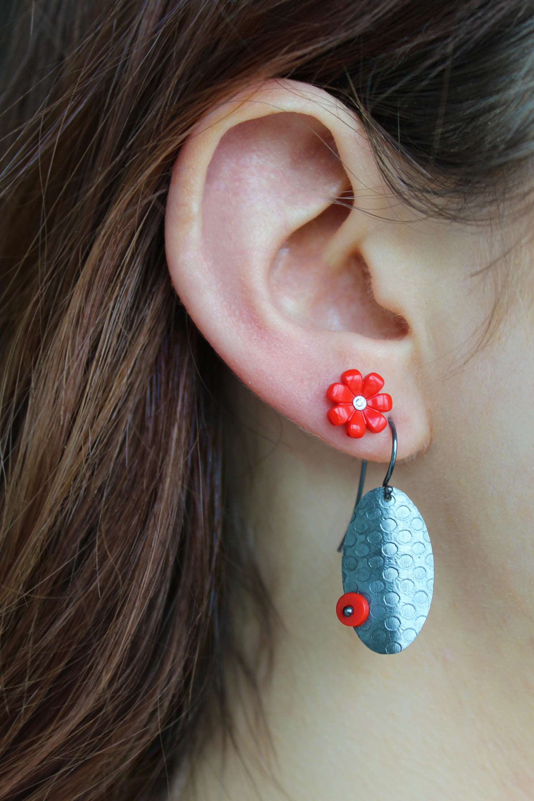 Two earrings on a womens ear. One being a litttle red daisy with a round diamond in the center and the other earring is an oval shape with a circle pattern on the metal. On one side, juxstapositioned is a red tiny circle riveted to towards bottom