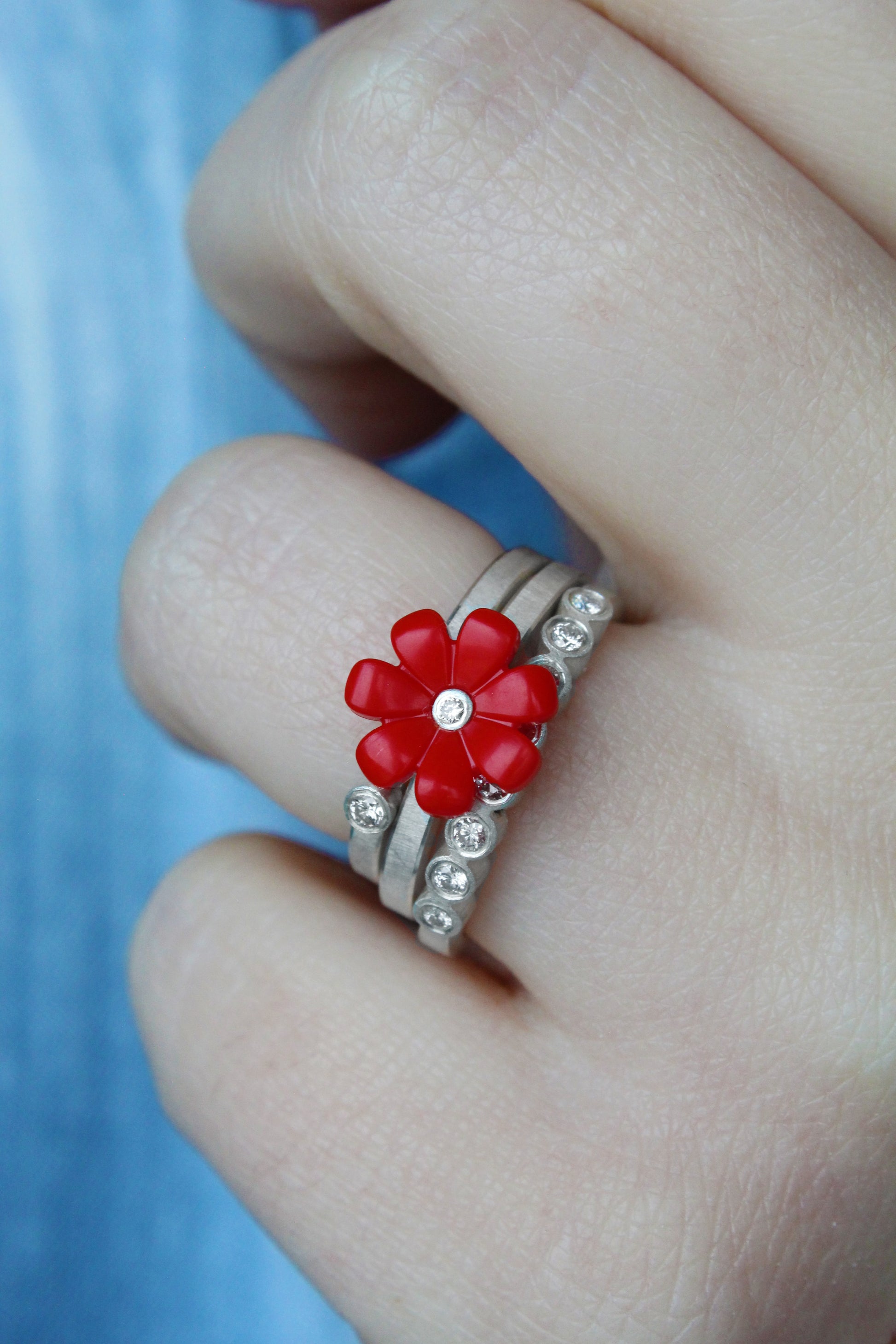 women displaying 3 sterling silver rings stacked on her finger. One has a red daisy with diamond in center, one is a band   sterling silver stackith one diamond set in a bezel and the other one has 8 diamonds going across on the top 