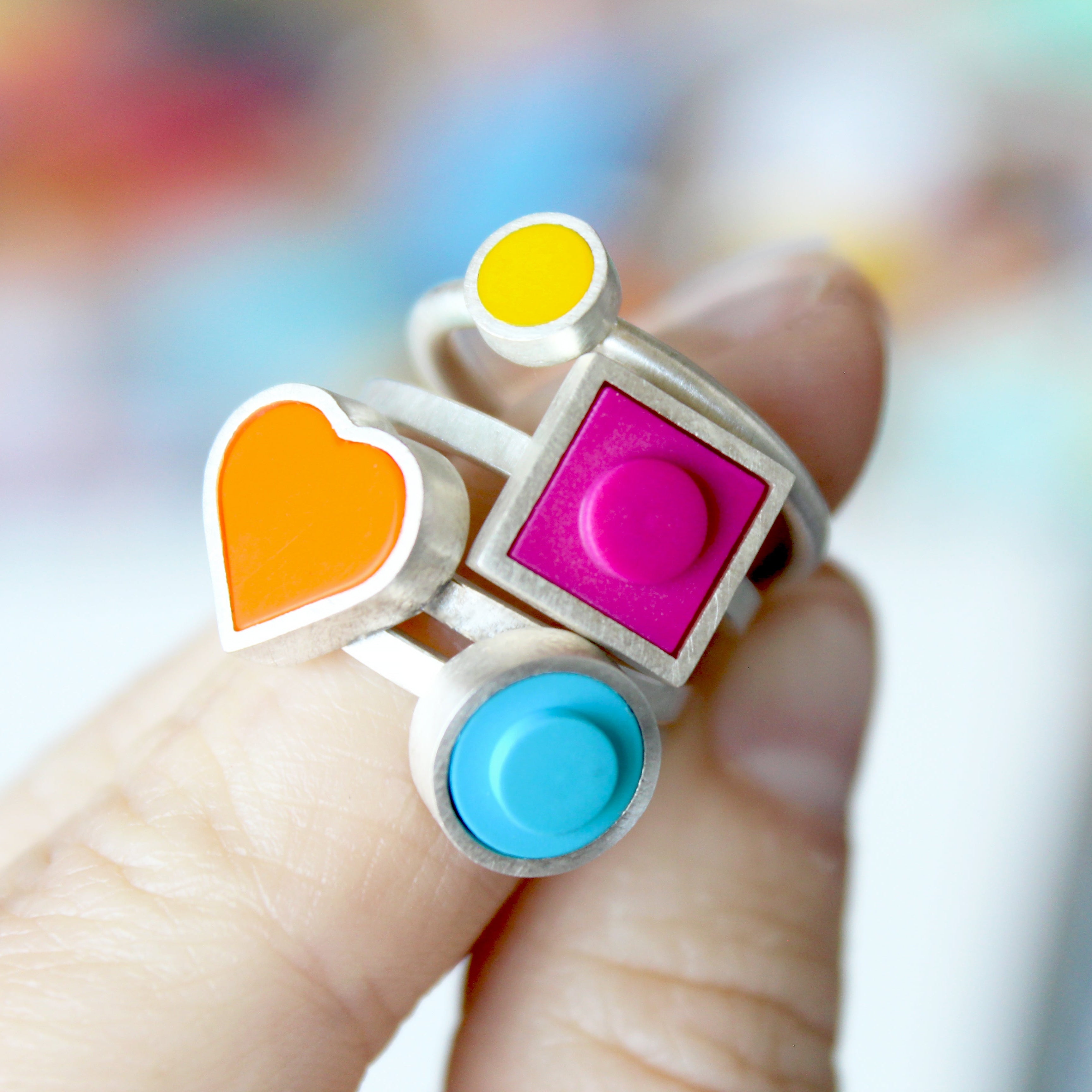 4 rings shown on stacked on a womens finger. Rings are, small yellow dot,  pink square, orange heart, and a blue round dot.